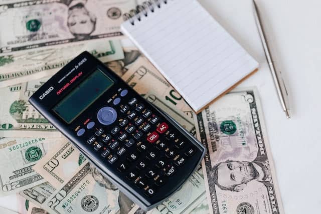 Calculator on top of dollar bills and notepad and pen next to them.